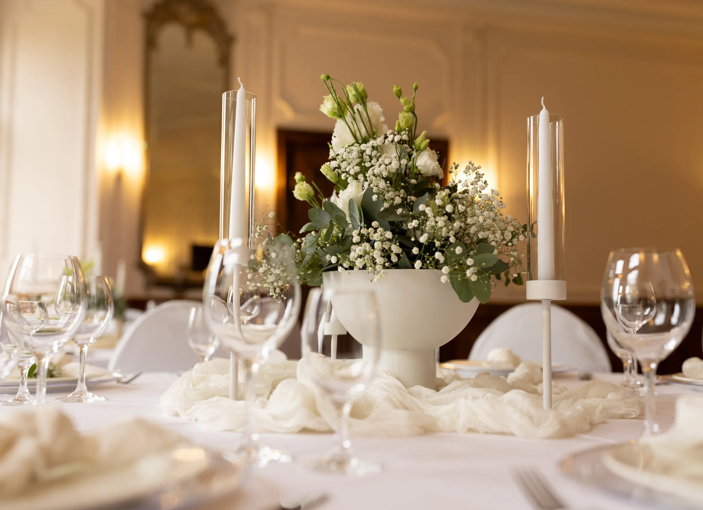 Festlich gedeckte Tafel mit weißem Blumenarrangement aus Rosen und Schleierkraut in einer Schale, Stabkerzen in Glaszylindern und Weingläsern